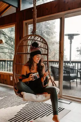 a woman sitting in a rattan chair holding a camera at Lightfoot Cabin Mid-Century Perfect Romantic Getaway Hot Tub Sauna in Running Springs
