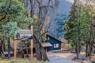 a sign in front of a blue house in the woods at River Haus Sequoia | Hot Tub+New+King Beds+Views! in Springville