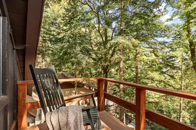 a rocking chair on a deck with a view of trees at Cute Romantic Cabin With Hot Tub 5 Min from Lake in Crestline
