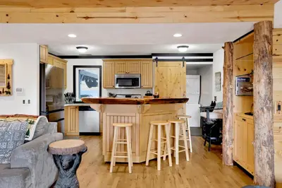 a kitchen and living room with wooden cabinets and a bar at Highland Hideaway at Big Bear | Cozy Cabin w/ Private Hot Tub in Big Bear Lake